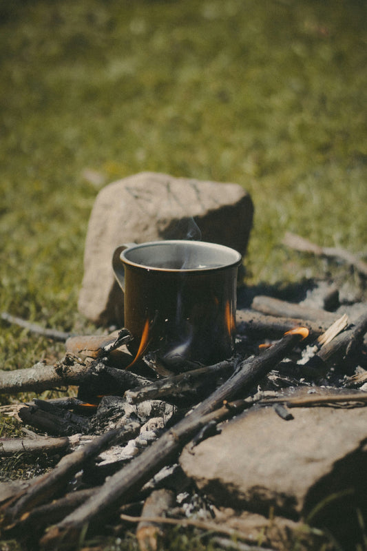 Metal camp mug heating over a small campfire made of sticks, with smoke rising and rocks surrounding the fire on a grassy outdoor campsite.