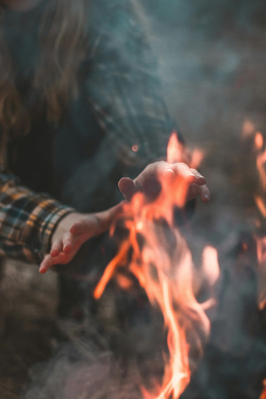 Person warming their hands over an open campfire, with visible flames and a flannel shirt, in a cold outdoor setting.