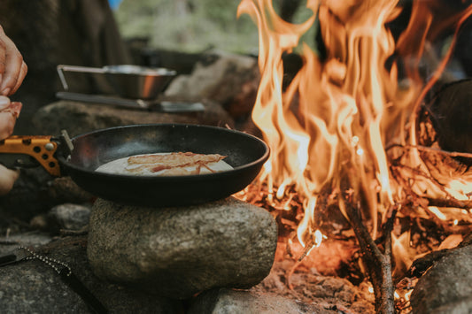 Bacon cooking in a pan set over rocks beside an open campfire, with visible flames and outdoor cookware in a rustic forest campsite