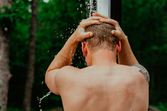 Man rinsing his hair under an outdoor shower surrounded by trees, with water streaming down his back in a forest setting.