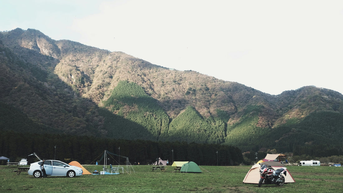 Scenic campground at the base of forested mountains with tents, a motorcycle, and cars set up on a grassy field under a bright sky.
