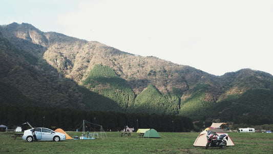 Scenic campground at the base of forested mountains with tents, a motorcycle, and cars set up on a grassy field under a bright sky.