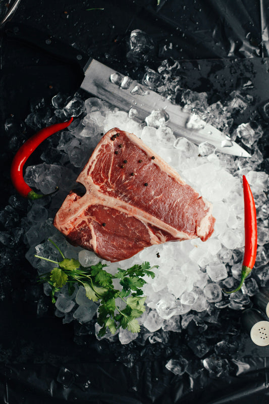 Raw steak with black peppercorns placed on a bed of ice, surrounded by red chili peppers, parsley, and a large chef’s knife on a dark surface.